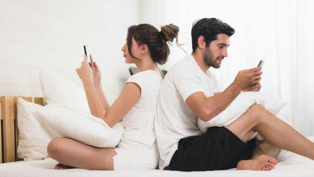 A man and woman sit back-to-back on a bed, both looking at their own smartphones.