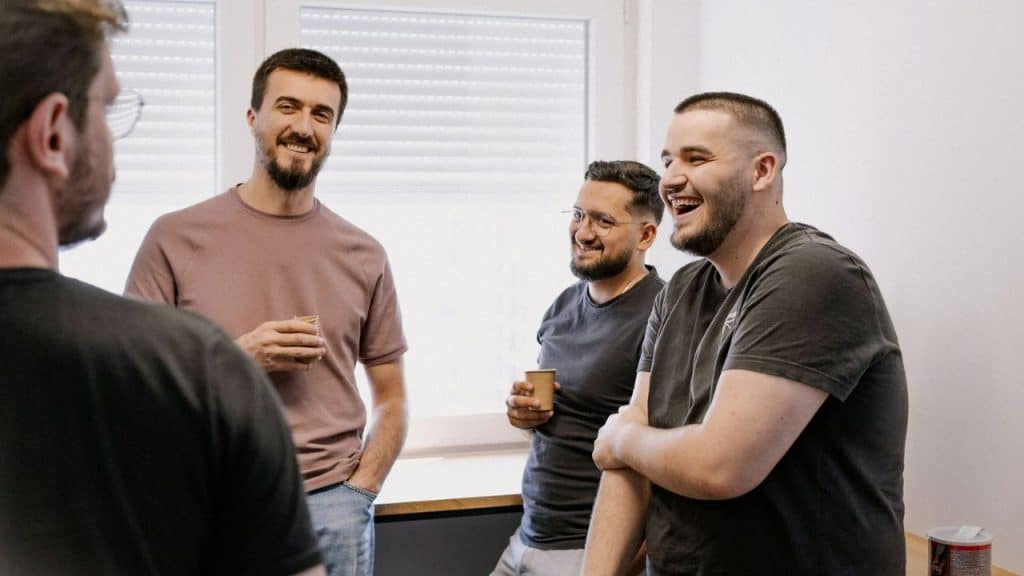 Three men stand in an office setting, laughing and holding small paper cups during a conversation.
