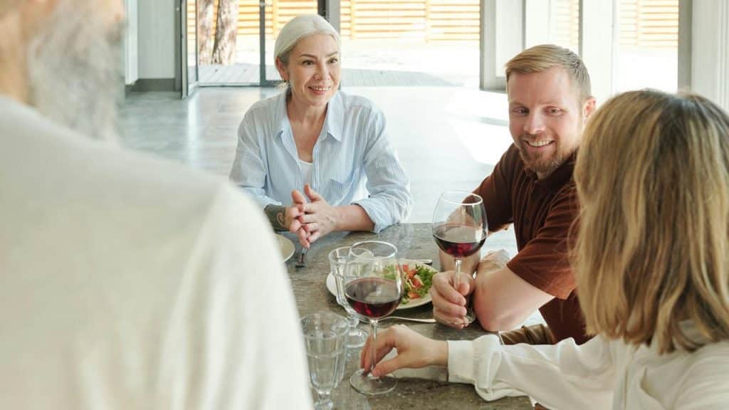A group of adults smiling and talking while sharing wine at a table indoors.