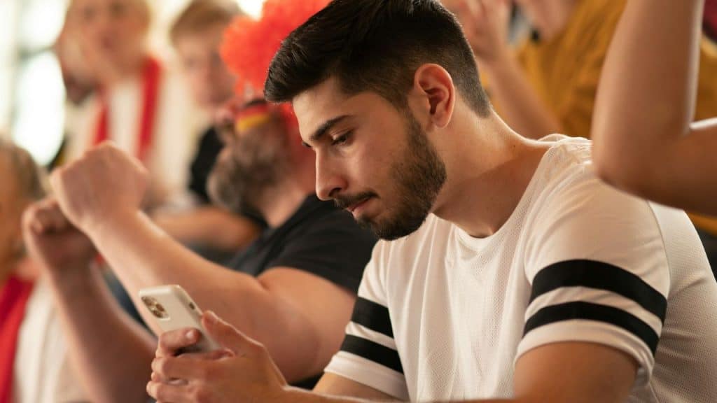 A man with a beard sits in a crowd and looks down at a white smartphone.