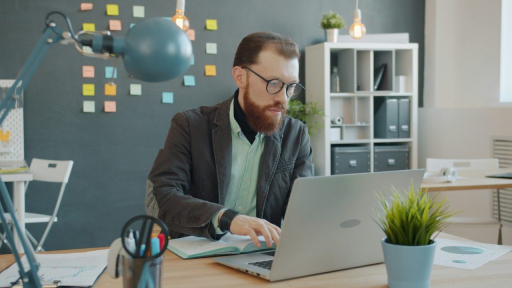 A man with a red beard wearing glasses works on a laptop at a desk.
