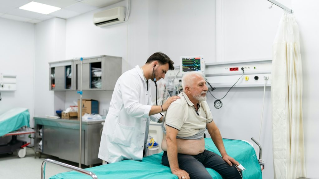 A doctor in a white coat uses a stethoscope on an elderly man's back.