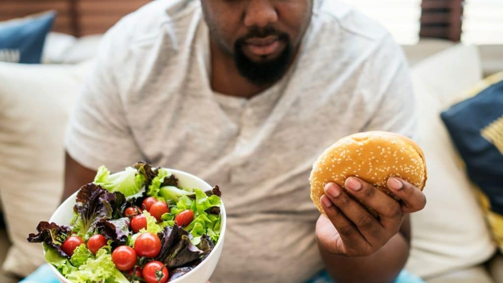 A man holds a fresh green salad in one hand and a sesame seed hamburger in the other.