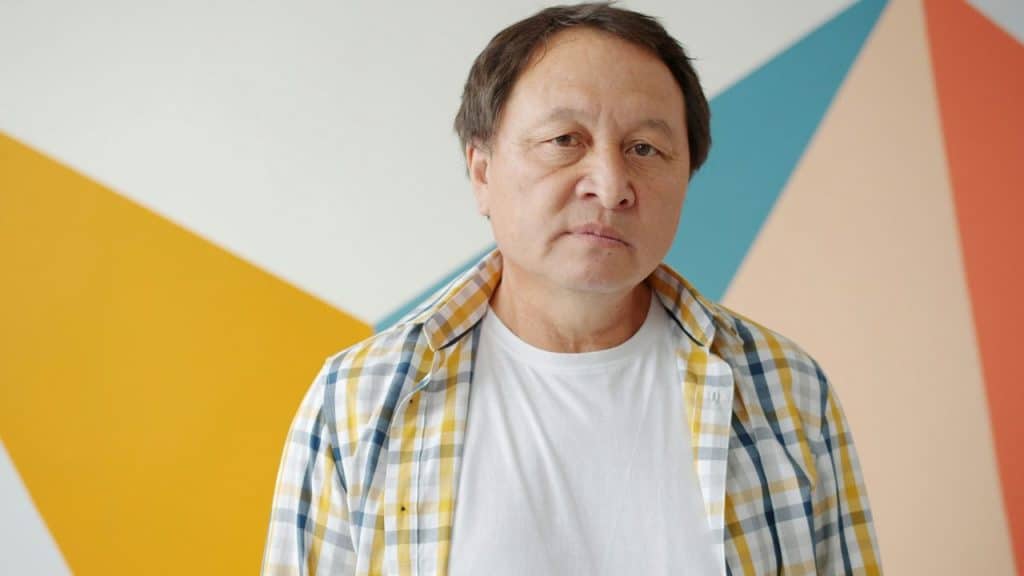A man in a yellow plaid shirt stands in front of a wall with geometric patterns.