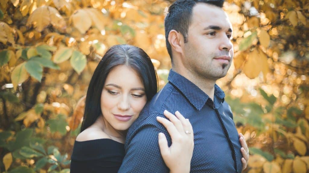 A woman hugging her husband from the backside while they are standing in autumn trees.
