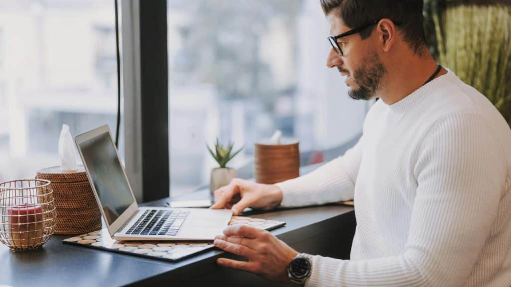 A man in glasses works on a laptop at a window-side desk.
