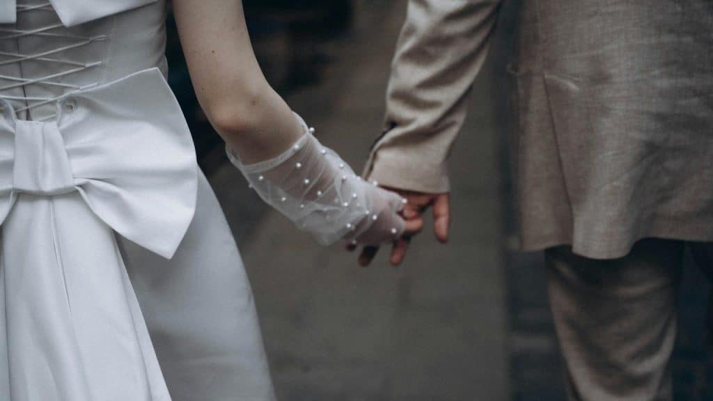 A bride in a white dress with a bow and a groom in a tan suit holding hands.