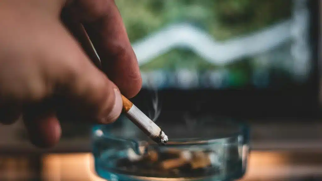 A hand holding a lit cigarette over an ashtray with a blurred line graph in the background.