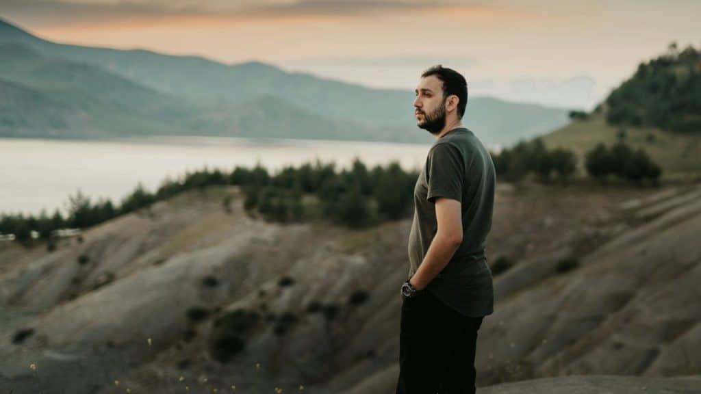 A bearded man in a dark t-shirt looks out over a valley and distant lake.