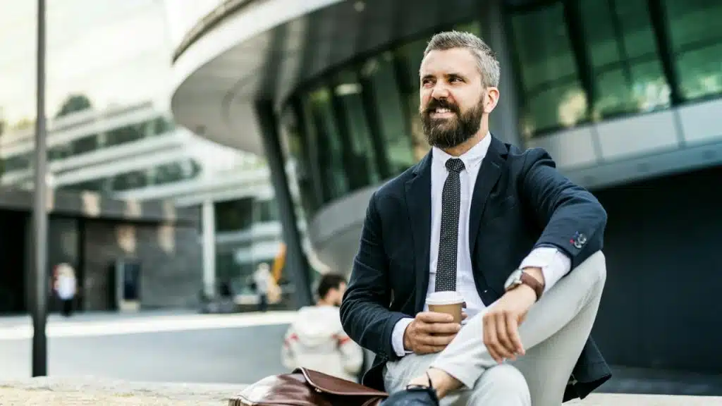 A bearded man in a suit sits outdoors holding a coffee cup and smiling thoughtfully.