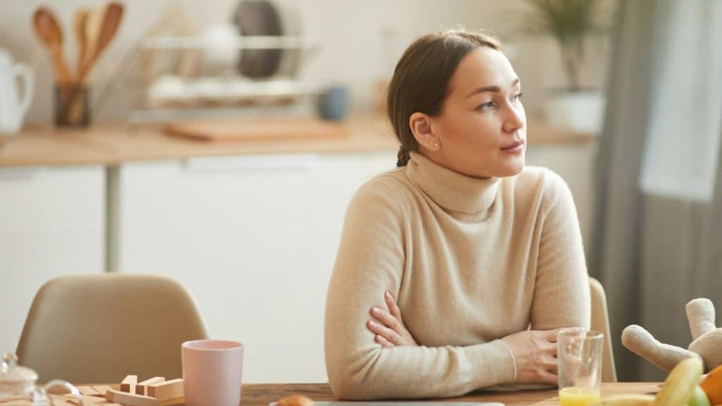 A woman in a turtleneck sweater sits at a table with her arms crossed, looking away.