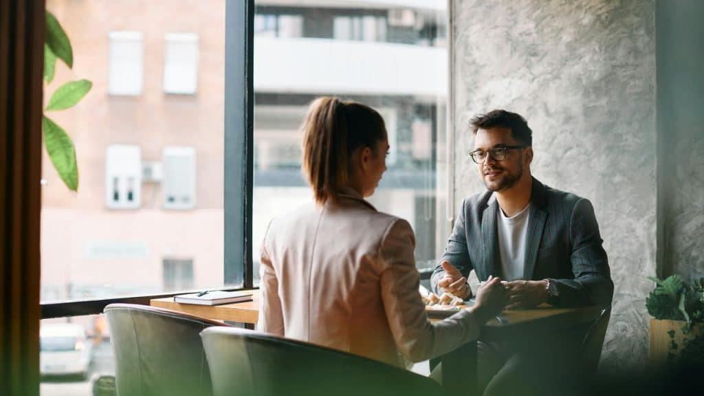 A man and woman talking over coffee by a window.