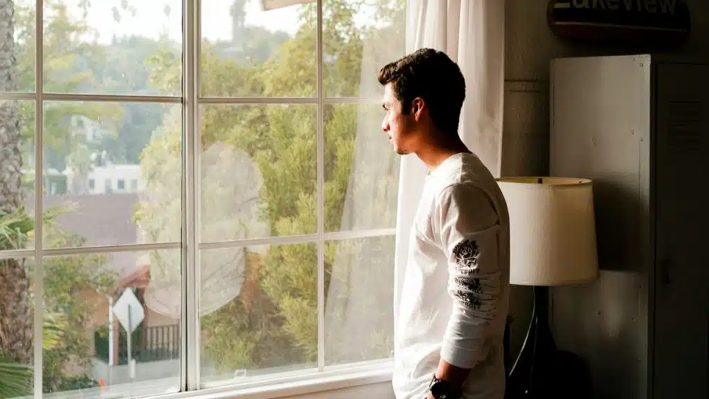 A man in a white long-sleeved shirt stands indoors and looks out of a large window.