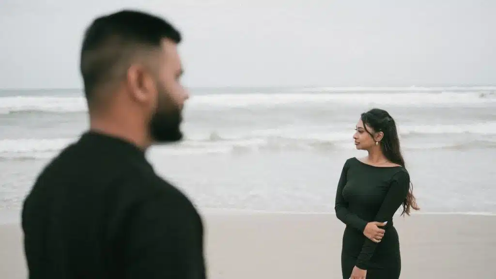 A bearded man looking to the side while a woman looks in the opposite direction as they stand on a beach.