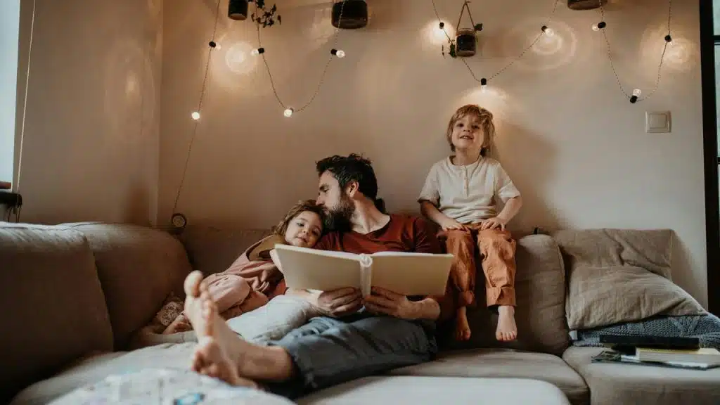 A parent reading a book with two young children on a couch under soft lights.