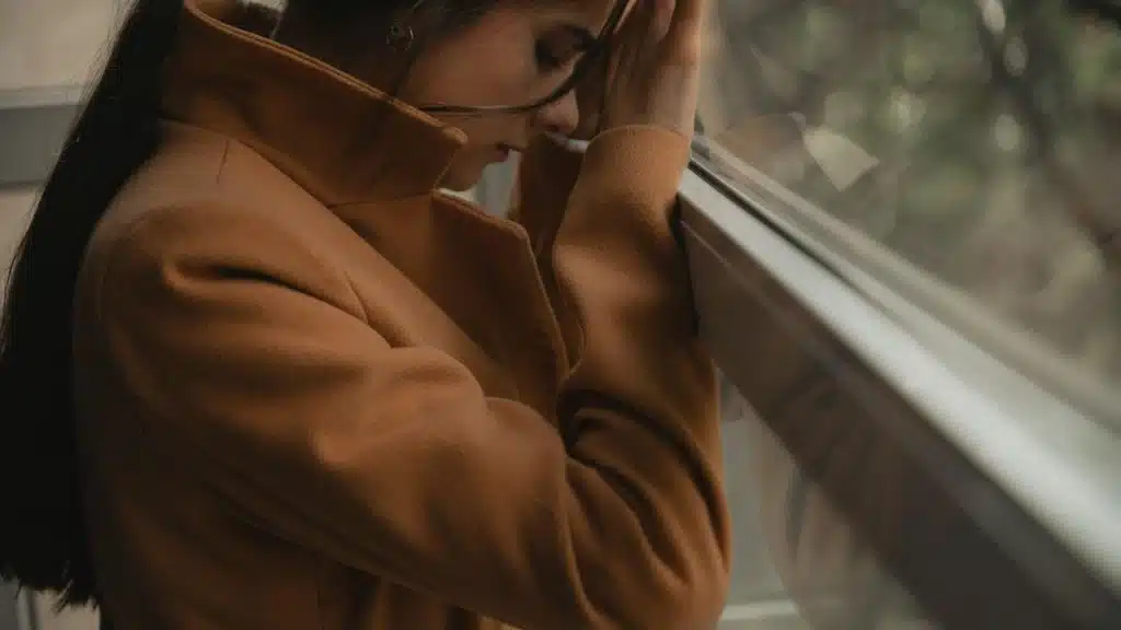 A person leaning against a window, resting their forehead on their hand.