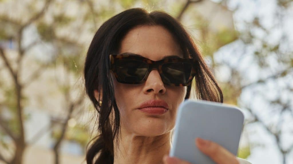 A woman wearing tortoiseshell sunglasses looks down at a light blue smartphone held in her hand.