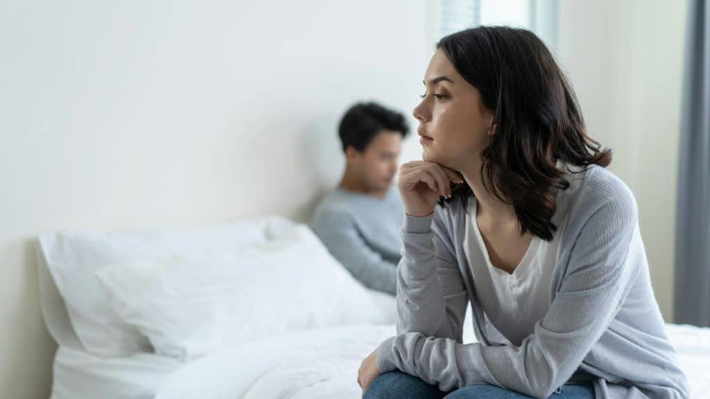 A woman with dark hair sits on a bed looking away from a man.