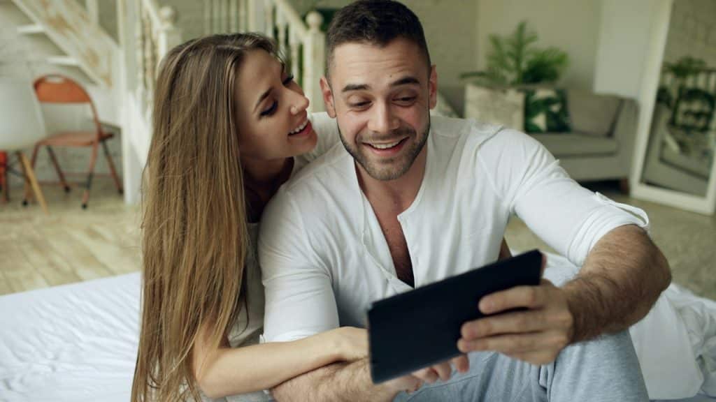 A woman leans against a man's shoulder as they both look at a tablet together.