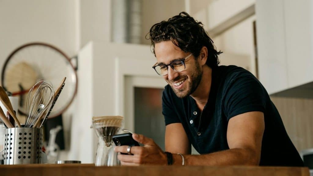 A smiling man with glasses leans on a kitchen counter while looking at his smartphone.
