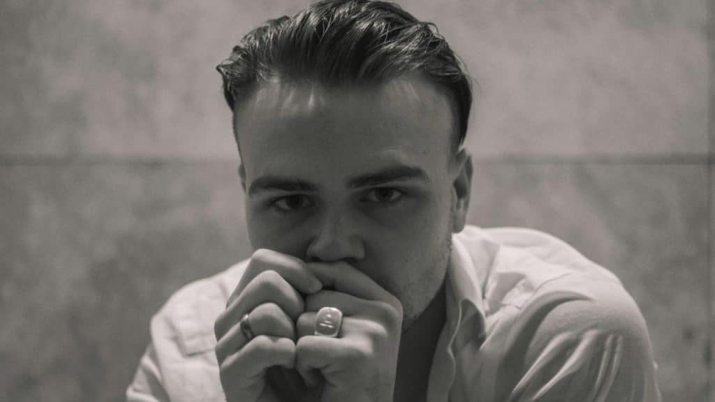 A man with slicked-back hair holds his hands to his mouth, looking directly forward.