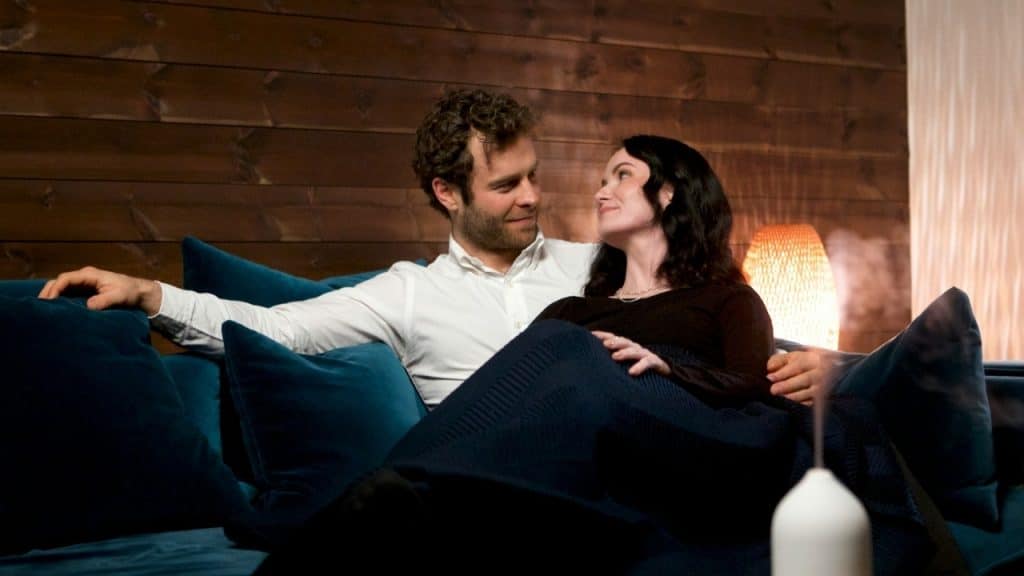 A man and woman sit closely together on a blue couch in a dimly lit room.