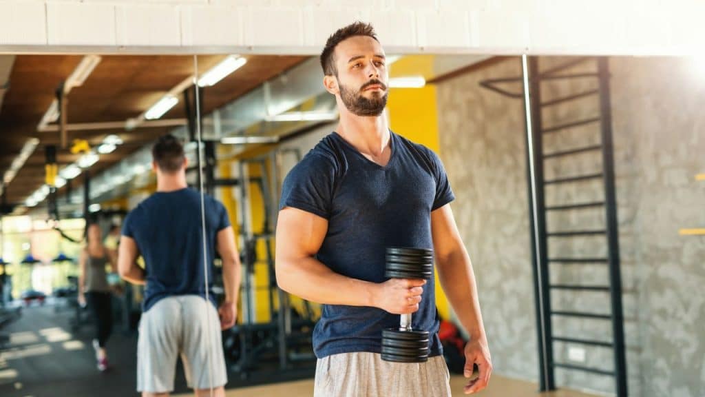 A man with a beard holds a dumbbell while looking into a gym mirror.