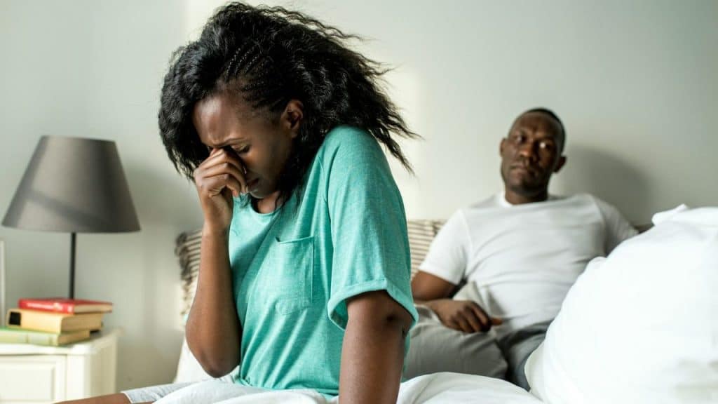 A woman sits on a bed covering her face while a man watches from behind.
