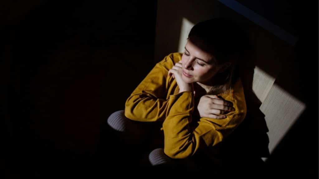 A woman in a yellow sweater sits on the floor in a beam of light.