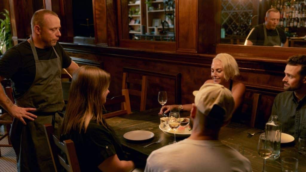 A server in a black apron stands by a wooden table where four people are seated.