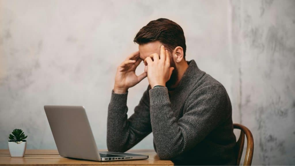 A man with a beard sits at a desk with a laptop, holding his head in his hands.