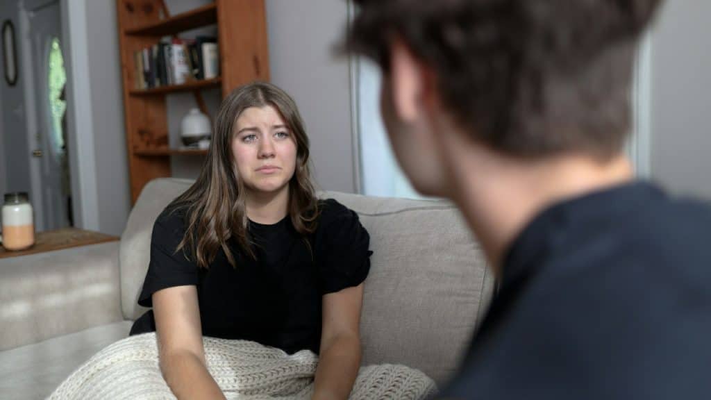 A woman with a sad expression sits on a couch looking toward a blurred person.