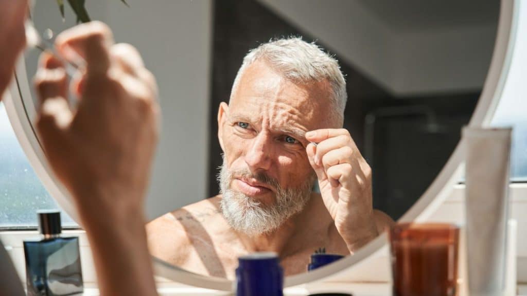 A man with a grey beard looks into a round mirror and tweezers his eyebrow.