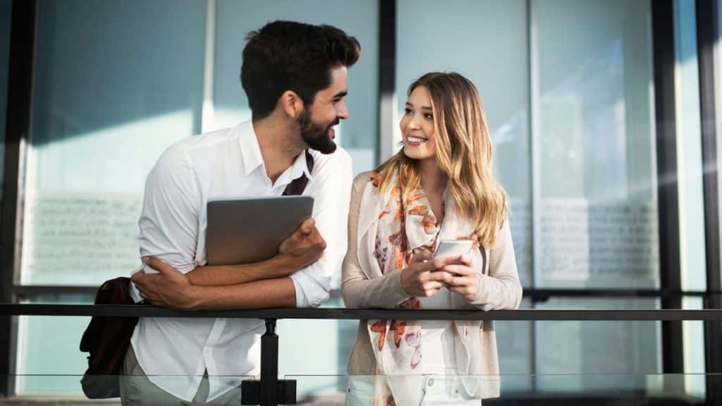 A man with a laptop and a woman with a phone smile while leaning on a railing.