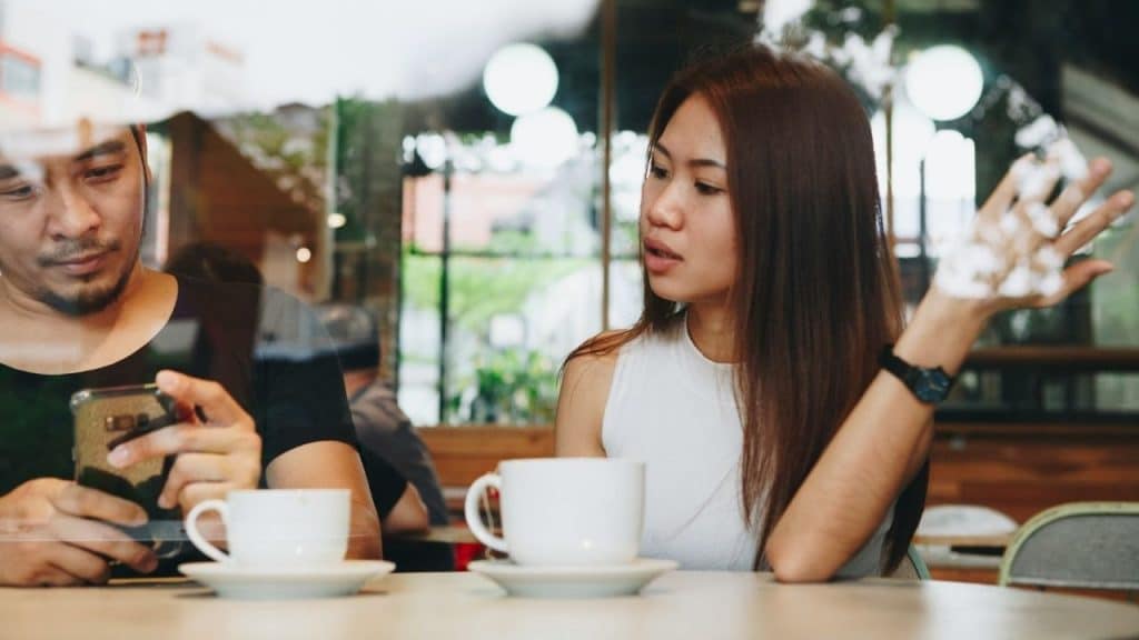 A woman talks and gestures while a man sits beside her looking at his phone.