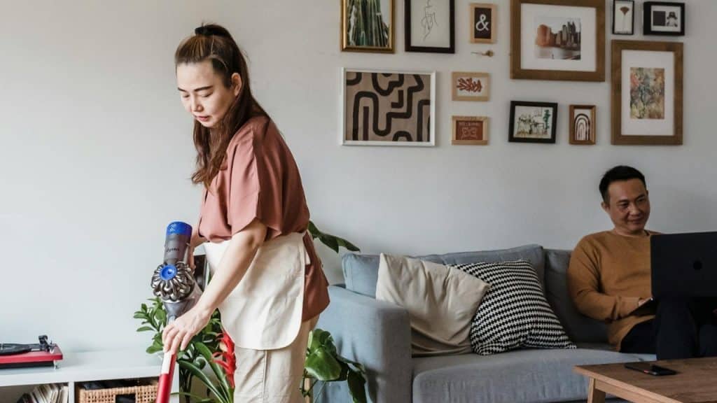 A woman vacuums a living room while a man sits on a couch using a laptop.