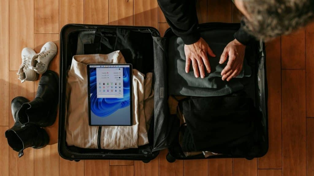 An overhead view shows a person's hands packing a tablet and clothes into a suitcase.