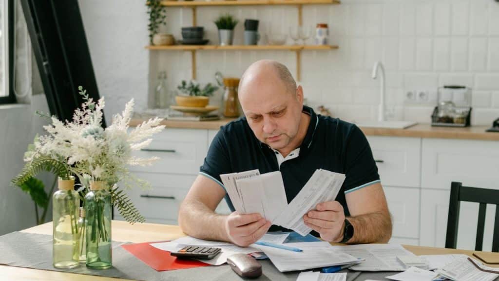 A bald man sits at a table in a kitchen, looking through several paper documents.