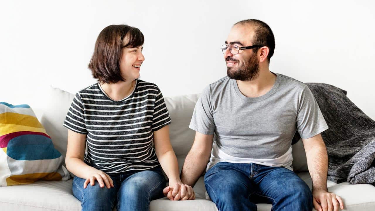 A casually dressed couple sits on a couch holding hands, smiling at each other.