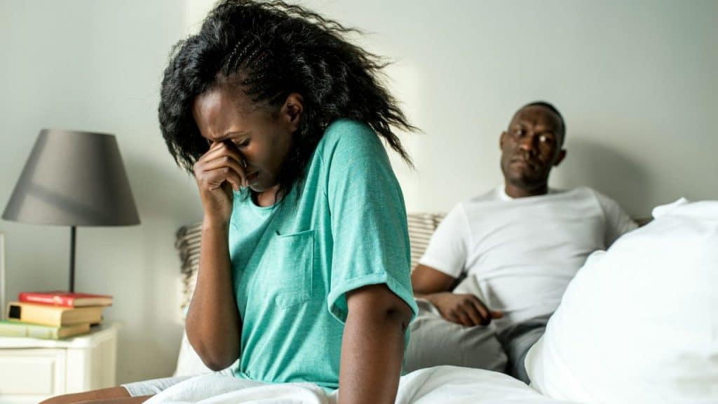 A woman sits on a bed crying while a man sits in the background looking away.
