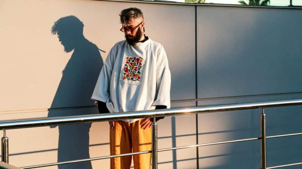 A bearded man wearing sunglasses and an oversized white shirt stands beside a metal railing.