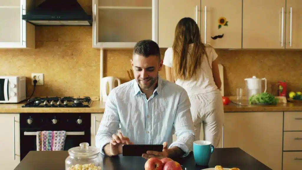 A man sits at a table using a tablet while a woman works in the kitchen.