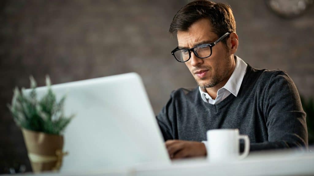 A man wearing glasses and a grey sweater looks intensely at a laptop screen.