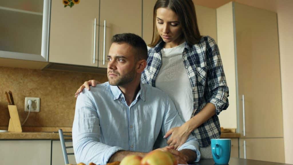 A woman stands behind a seated man in a kitchen, placing her hands on his shoulders.