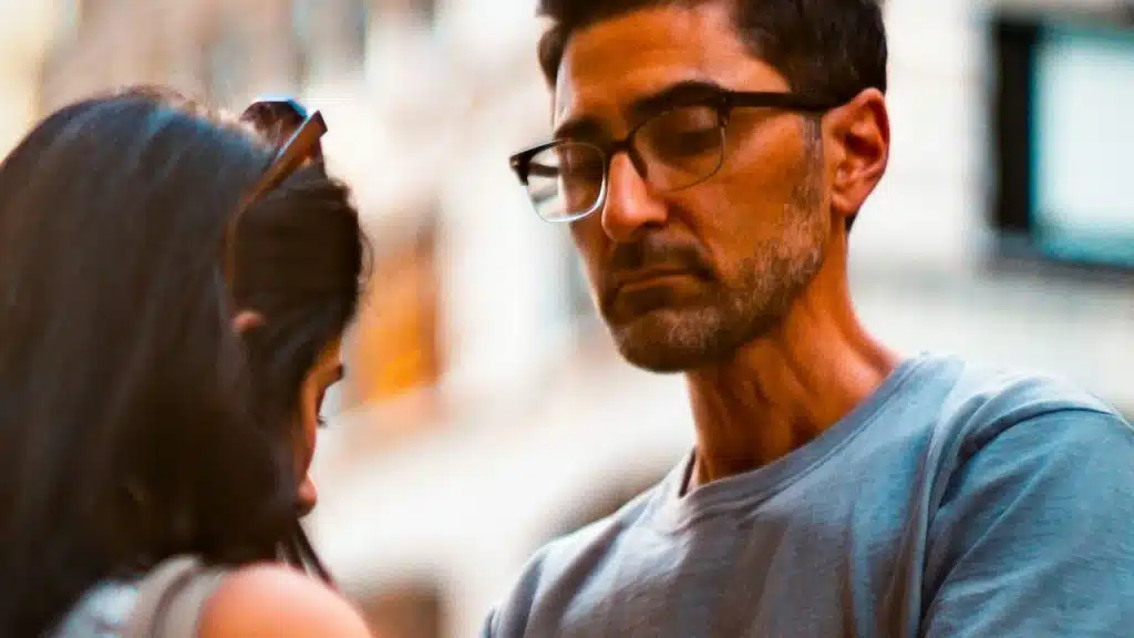 A man wearing glasses looking upset and looking away while a woman stands in front of him and looks downward.