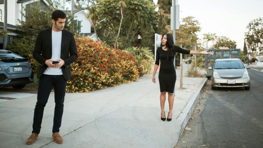 A woman gestures on a sidewalk while a man nearby looks down at his phone.