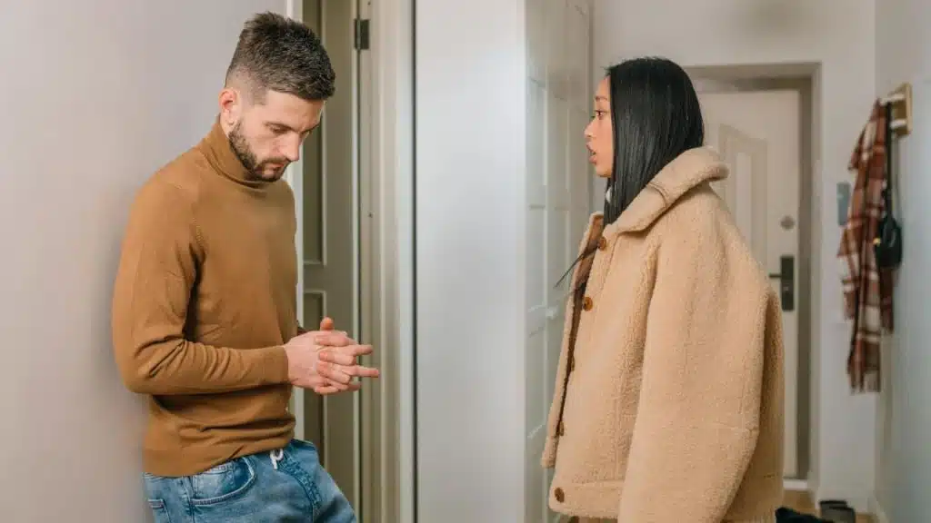 A man leans against a wall looking down while a woman in a coat faces him.