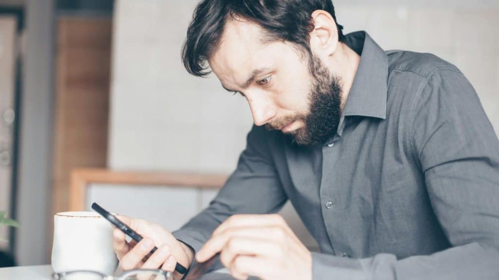A bearded man in a grey button-down shirt looks down intently at a smartphone.
