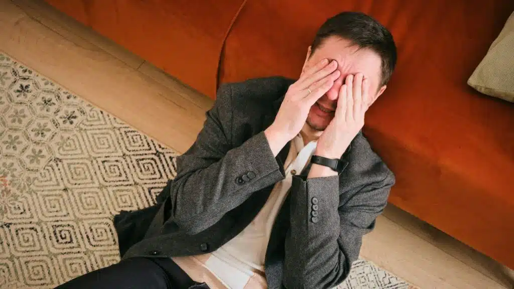 A man in a blazer sits on a rug covering his face with both hands.