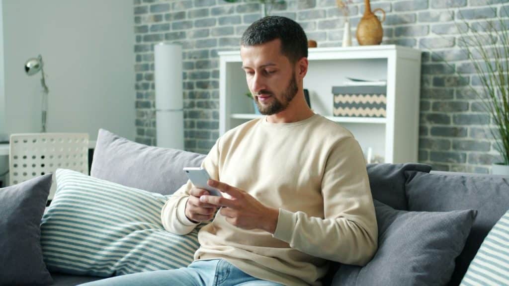 A bearded man in a beige sweater sits on a gray couch looking at a smartphone.